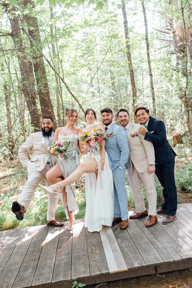 A joyful wedding party poses together on a wooden path in a forest, smiling and laughing. The bride, groom, and attendants hold colorful bouquets, with some playfully kicking up their legs. Sunlight filters through the trees.