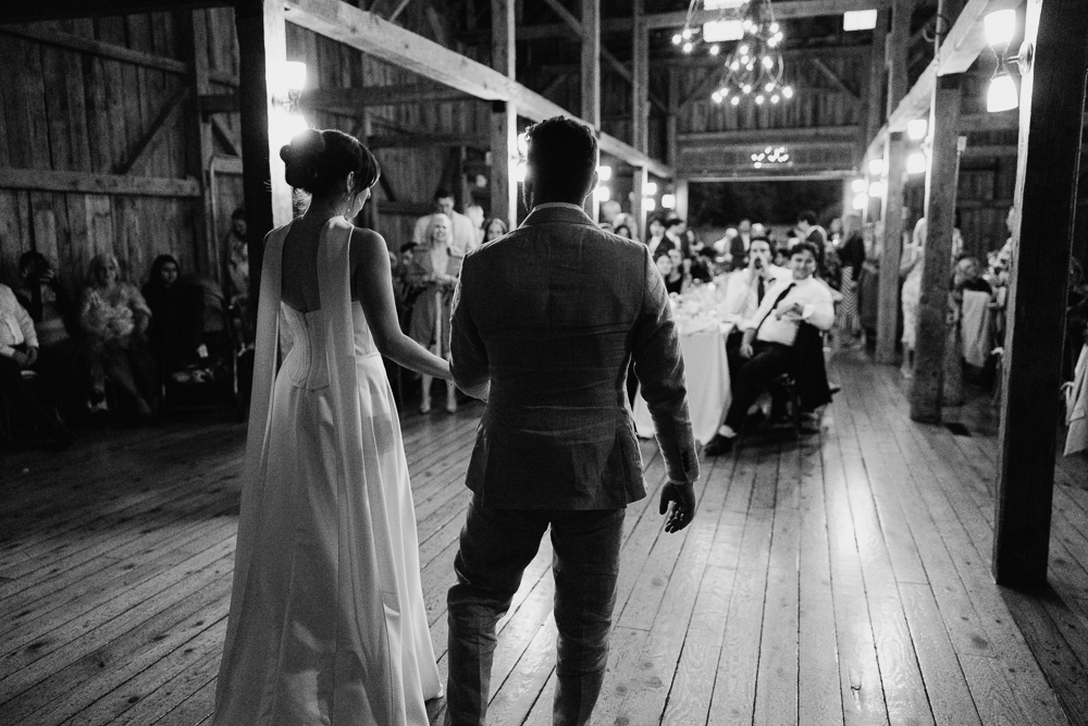 A bride and groom, seen from behind, walk hand in hand into a rustic barn venue with wooden floors, where guests are seated and watching them. The scene is warmly lit and festive.