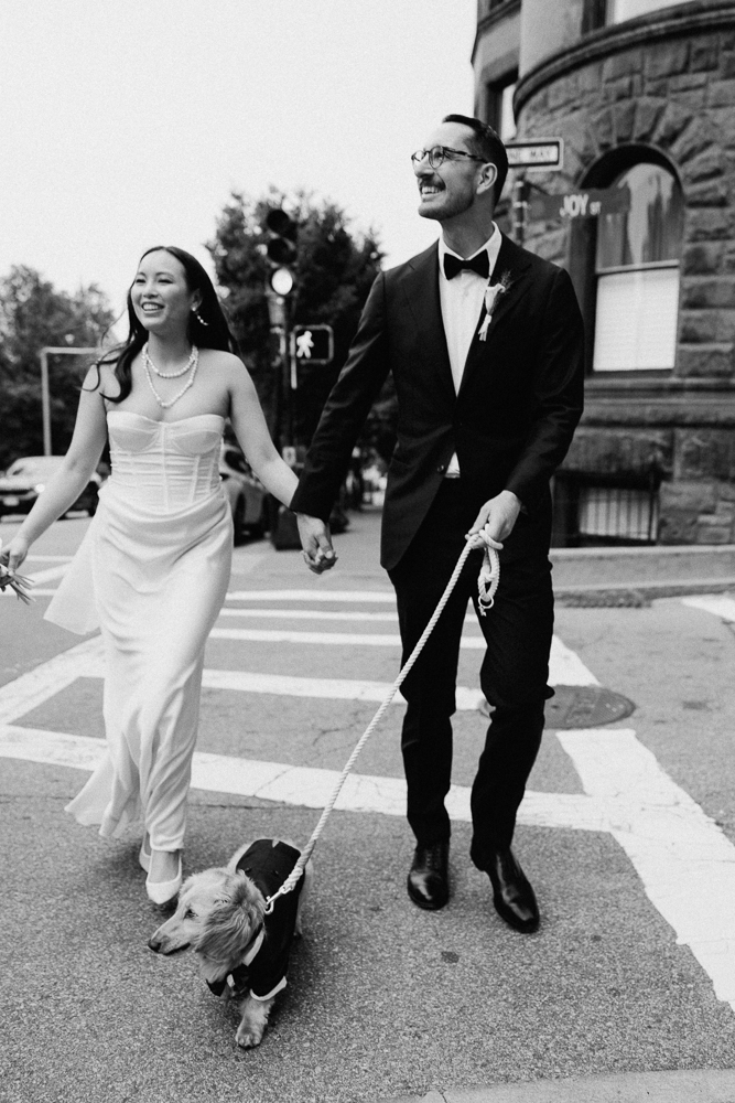 A bride and groom, both smiling and holding hands, walk across a street with their dog dressed in a tiny tuxedo. The image is black and white, and they appear joyful and celebratory.