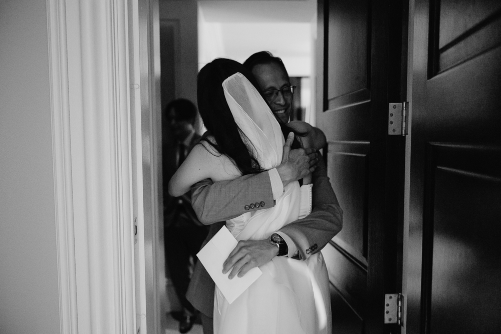A bride in a wedding dress and veil hugs a man wearing glasses and a suit in a hallway. He smiles warmly and holds an envelope in one hand. The moment appears candid and joyful.