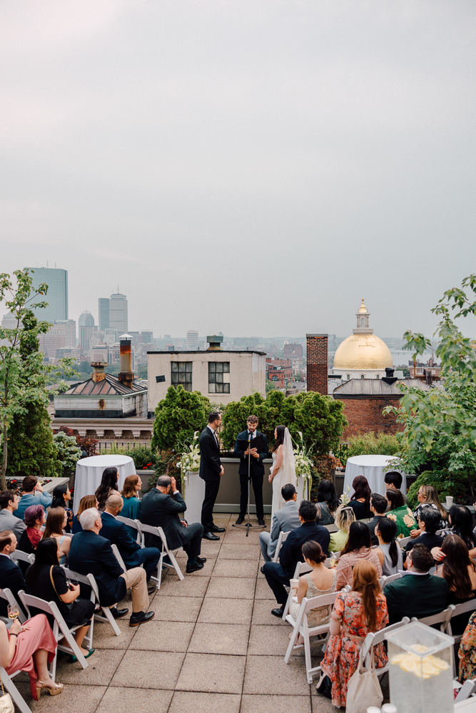 A rooftop wedding ceremony with a city skyline in the background; guests are seated facing a couple and officiant under a decorated arch, with greenery and a gold-domed building visible behind them.