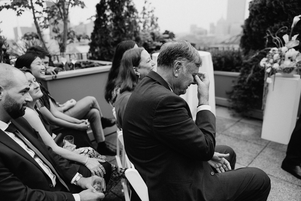 A group of people sit in rows at an outdoor event. One older man in a suit wipes his eyes, appearing emotional, while others around him smile. There are plants and city buildings in the background.