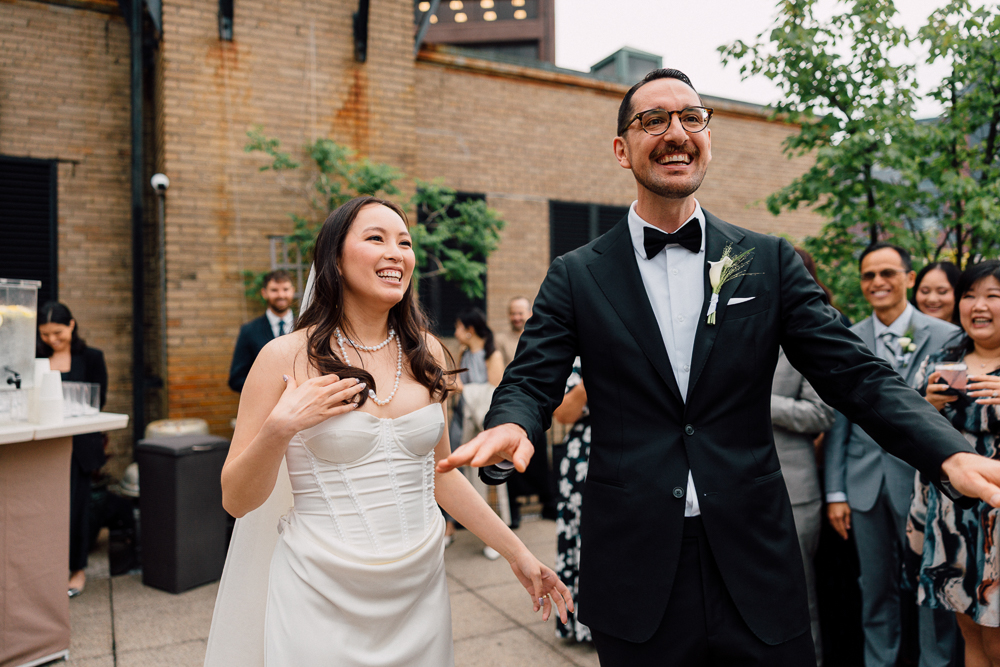 A smiling bride in a white dress and a groom in a black tuxedo stand together outdoors, surrounded by joyful guests at a wedding celebration.