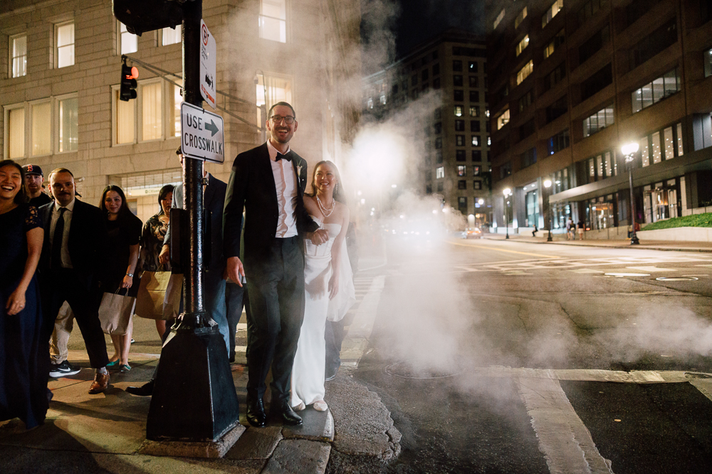 A bride and groom stand on a city street at night near a crosswalk, surrounded by friends, with steam rising from a grate nearby and tall buildings in the background.