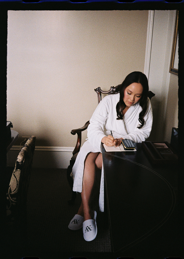 A woman in a white bathrobe and slippers sits at a desk, writing in a notebook while using a calculator. She appears focused, seated in a chair in a softly lit room with beige walls and carpet.