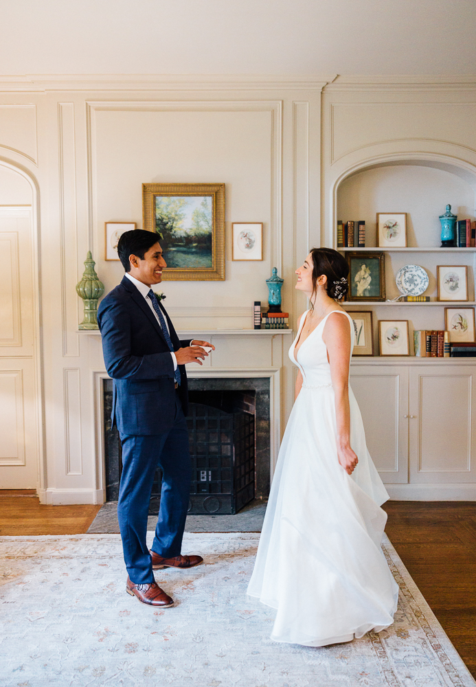 A bride in a white gown and a groom in a navy suit stand facing each other, smiling, in a bright, elegantly decorated room with bookshelves, framed art, and decorative vases—capturing the charm of Moraine Farm Wedding Photography.