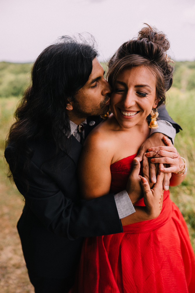 A man in a dark suit embraces and kisses a smiling woman in a red dress and gold earrings outdoors. The couple appears happy and affectionate, standing on a path with greenery—a moment perfect for Moraine Farm Wedding Photography.