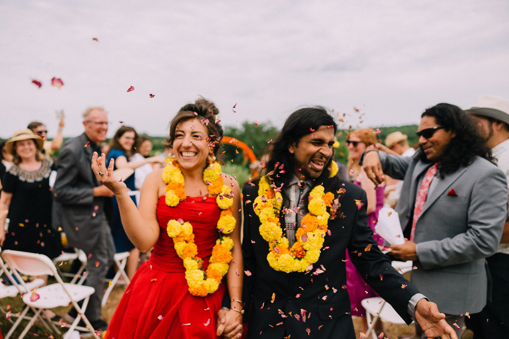A joyful couple, wearing large yellow flower garlands, walks hand in hand down an aisle outdoors as guests toss flower petals and cheer. The woman wears a red dress, the man a dark suit, both smiling brightly.