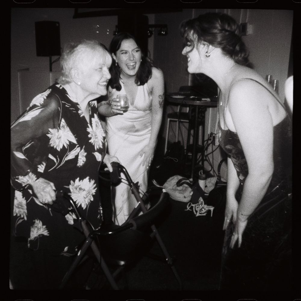 Three women laugh and interact at a lively indoor event. One older woman uses a walker, while the others, both in elegant dresses, smile and lean in towards her. The atmosphere is joyful and celebratory.