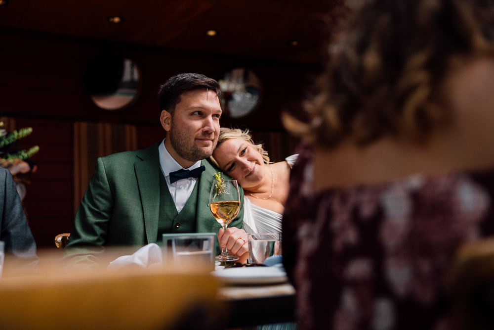 A man in a green suit sits at a table holding a wine glass, while a woman in white leans affectionately on his shoulder, smiling. The scene appears warm and intimate, with other guests blurred in the foreground.