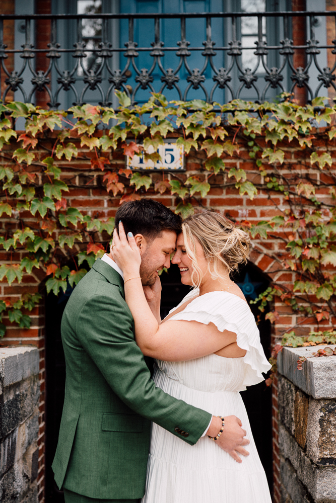 A couple embraces and smiles, touching foreheads in front of a brick building covered in green and red ivy. The man wears a green suit, and the woman wears a white dress.