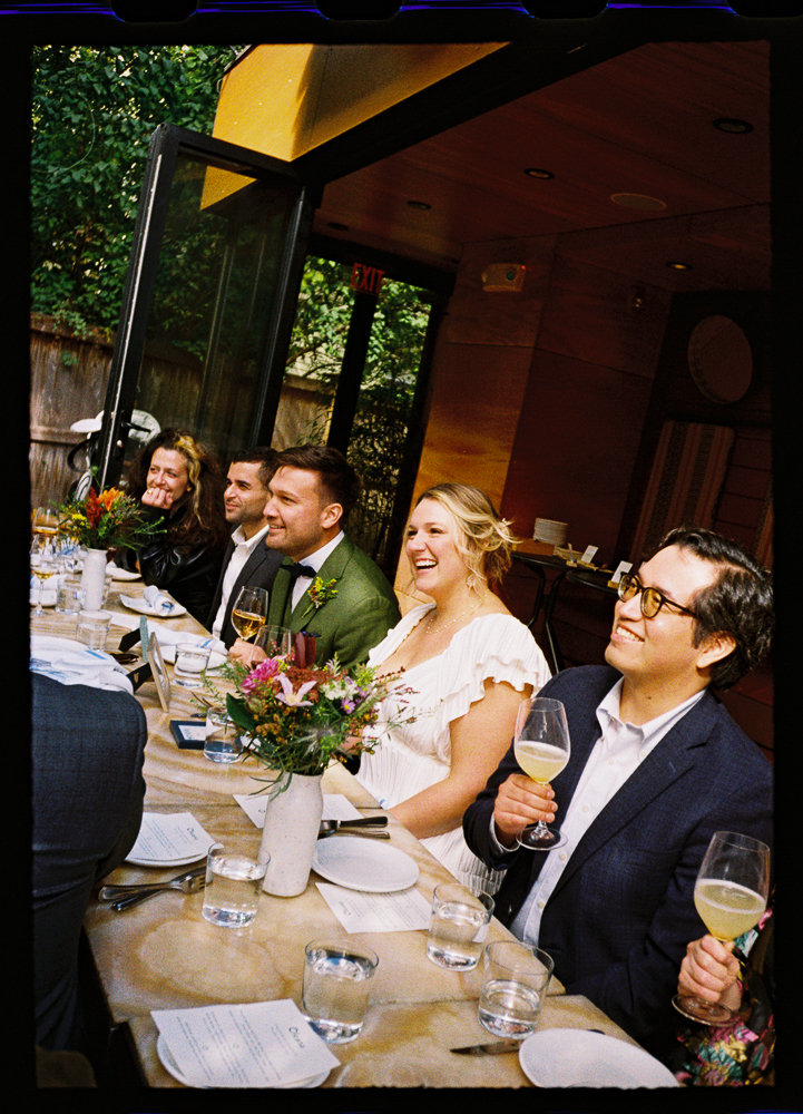 A group of people, including a bride in a white dress and a groom in a green suit, sit at a decorated table indoors, smiling and holding glasses during a celebratory meal.