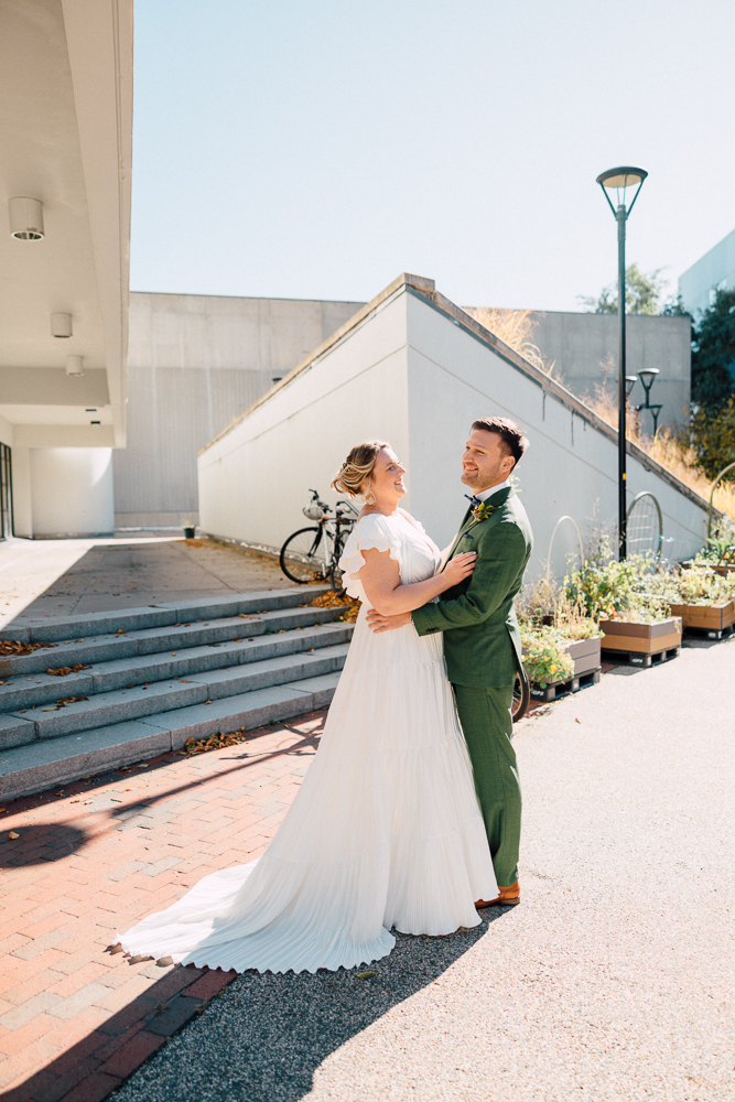 A bride in a flowing white dress and a groom in a green suit smile and embrace outdoors on a sunny day, standing near steps and a bike, with modern buildings and garden planters in the background.