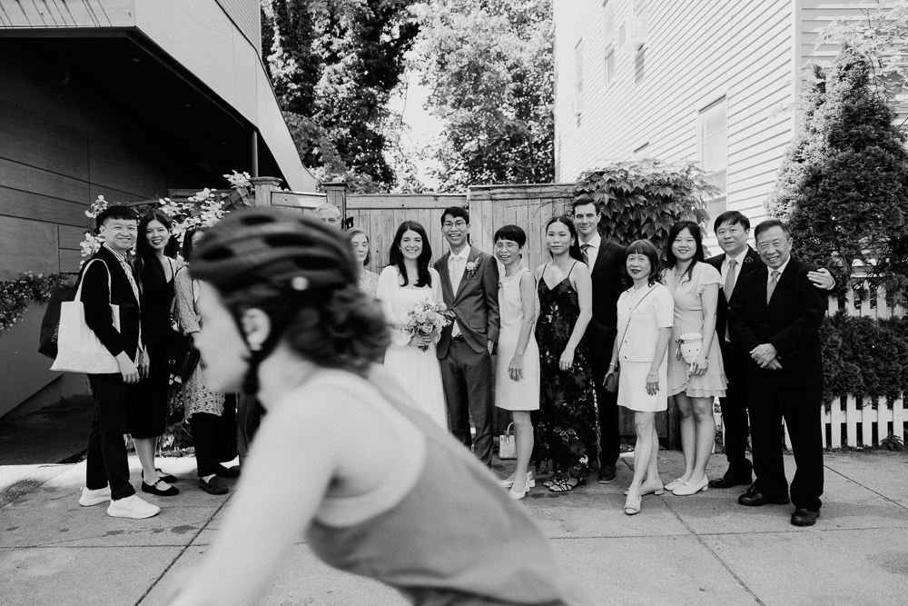 A group of people dressed for a wedding poses and smiles outside, while a person wearing a bike helmet rides past in the foreground, slightly blurred. The scene is in black and white.