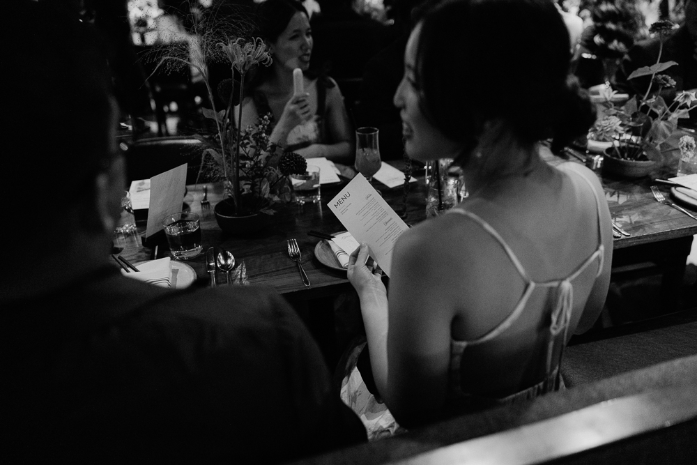 A woman in a sleeveless dress holds a menu while seated at a restaurant table, surrounded by food, drinks, and other diners in a warmly lit, lively atmosphere.