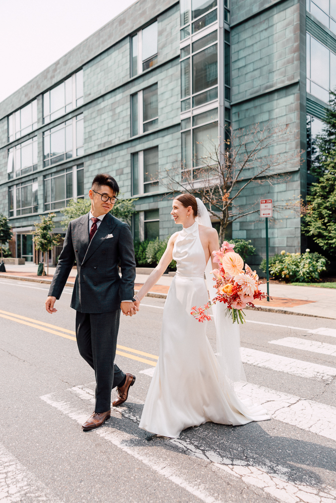 A bride in a white dress and groom in a dark suit walk hand in hand across a city street, smiling at each other. The bride holds a large, colorful bouquet; modern buildings are in the background.