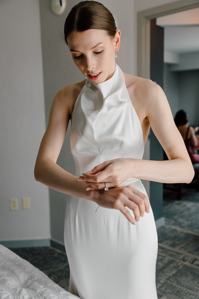 A woman in a white sleeveless halter dress adjusts a bracelet on her wrist while standing indoors. She has light skin, brown hair pulled back, and appears to be getting ready, possibly for a special event.