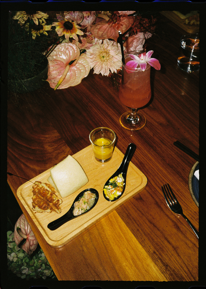 A wooden table with a wooden platter holding a bread roll, waffle piece, two appetizer spoons, and a shot glass of soup. A pink cocktail with an orchid garnish and flowers are in the background.