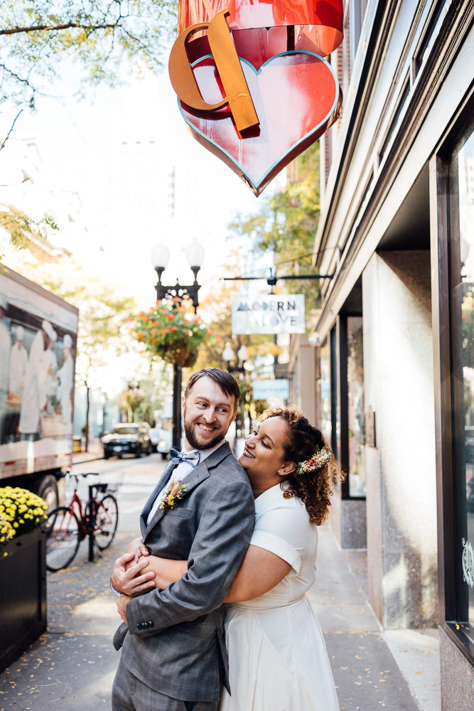A joyful couple in wedding attire embraces on a city sidewalk, smiling and laughing under a large red heart sculpture. Bicycles, flowers, and a bus are visible in the background on a sunny day.