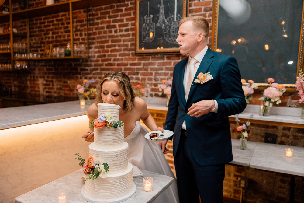 A bride leans forward to take a bite out of a white three-tiered wedding cake with flowers, while a groom in a suit stands nearby, smiling with cake on his mouth and holding a plate and fork.