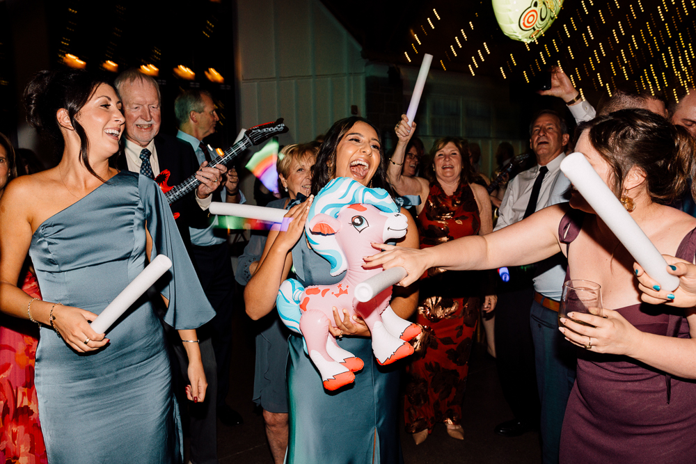 A group of people dance and laugh at a lively party. A woman in the center, holding an inflatable unicorn, smiles joyfully. Others around her hold glow sticks and drinks, celebrating under festive lights.