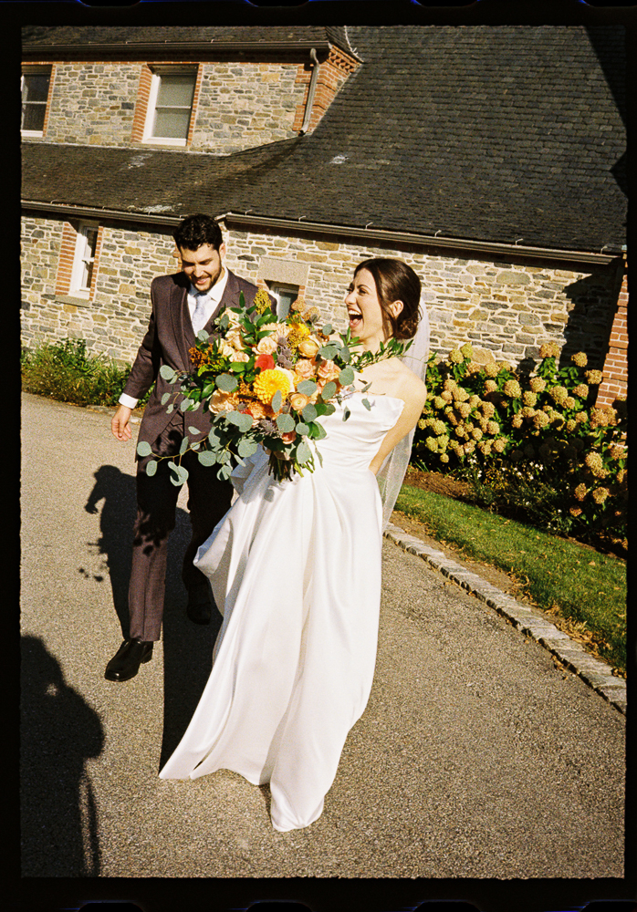 A joyful bride in a white dress holding a large, colorful bouquet walks outdoors with a smiling groom in a dark suit, in front of a stone building with flowers and greenery.