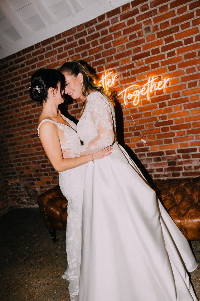 Two brides in white dresses embrace and smile with their foreheads touching in front of a brick wall and a neon sign that reads “Better Together.”.