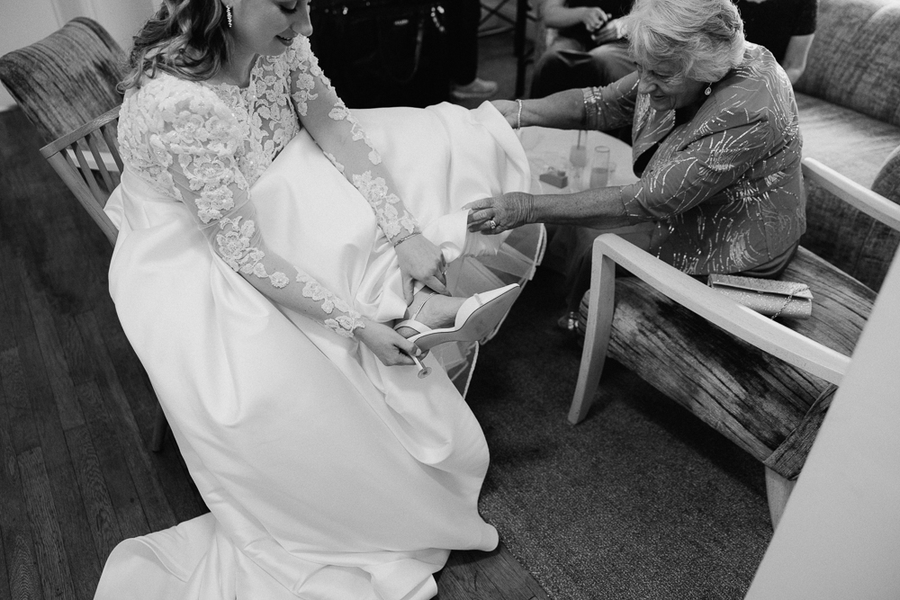 A bride in a lace wedding dress sits and puts on shoes, while an elderly woman in a patterned dress helps her. They are indoors, sharing a joyful, intimate moment together.