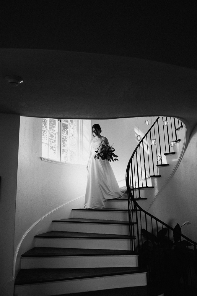 A bride in a white gown holding a bouquet descends a curved staircase, illuminated by natural light from a window. The scene is in black and white, creating a classic, elegant atmosphere.
