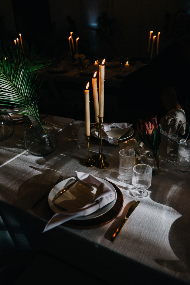 A dimly lit, elegant dinner table set with white plates, folded napkins, gold cutlery, glassware, and tall lit candles. Palm leaves and flowers in clear vases serve as centerpieces.