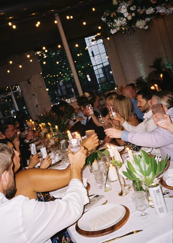 A group of people sitting around a long, decorated table raise their glasses for a toast at a festive indoor event with string lights and floral centerpieces.