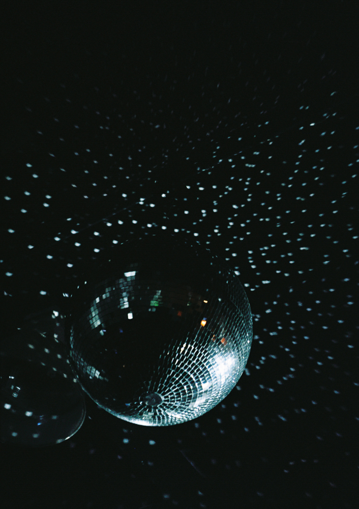 A disco ball hangs in the dark, reflecting light and creating a pattern of small, scattered white dots across the surrounding black space.