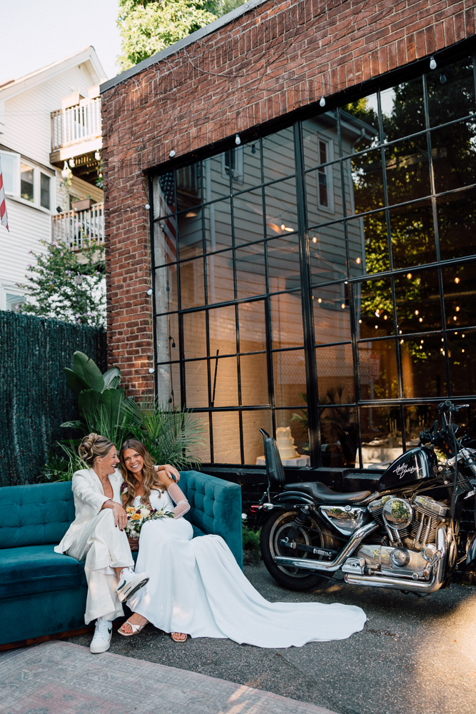 Two brides in wedding attire sit together on a blue couch outside a brick building with large windows. They smile at each other, holding a bouquet, with a black motorcycle parked nearby.