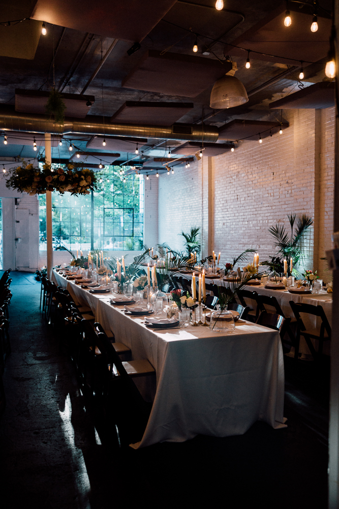Elegant indoor dining setup with long tables covered in white tablecloths, decorated with candles, glassware, and flowers. String lights hang overhead, and large windows let in natural light.