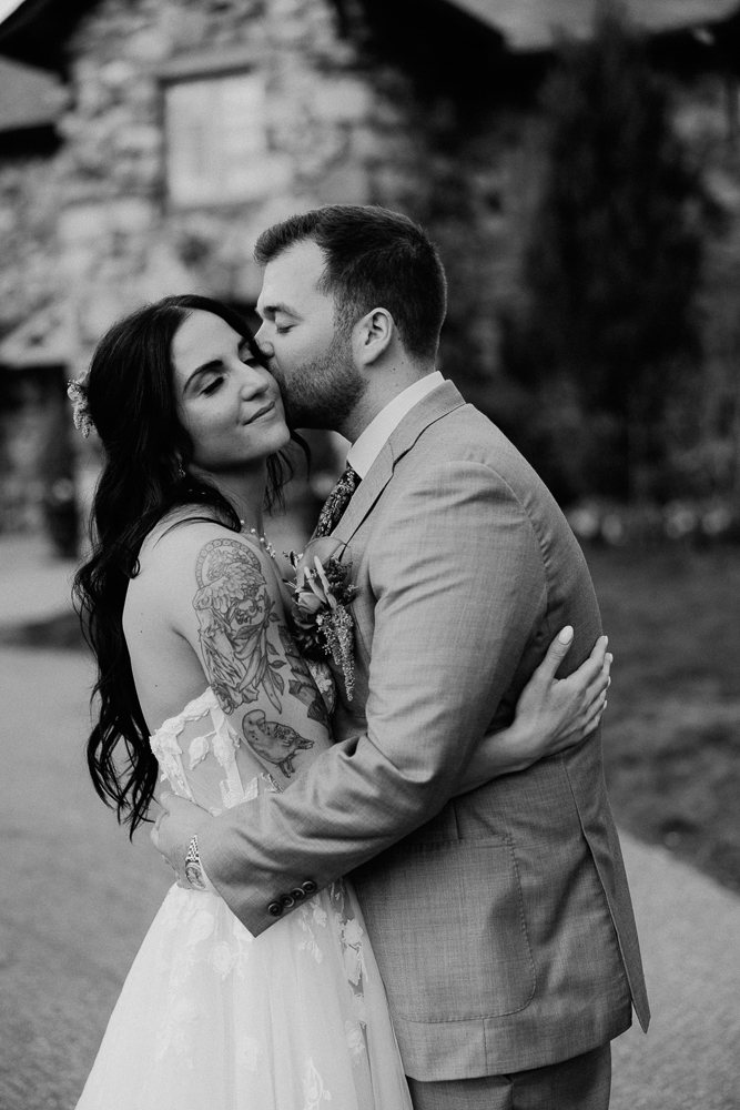 A bride with long dark hair and visible tattoos smiles with her eyes closed as a groom in a suit embraces and kisses her forehead. They stand outdoors in front of a stone building. The photo is in black and white.