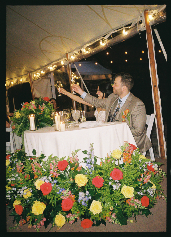 A bride and groom sit at a decorated table under a tent with string lights, raising their glasses in a toast. The table and front area are adorned with large, colorful flower arrangements.