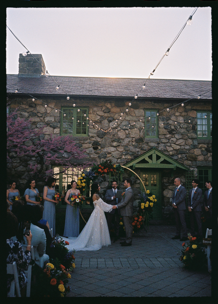 A wedding ceremony outdoors in front of a rustic stone building. The bride and groom stand under a circular floral arch, surrounded by bridesmaids in blue dresses and groomsmen in gray suits. String lights hang above.