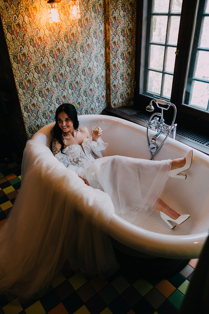 A bride in a white gown and heels lounges playfully in a vintage bathtub, smiling and holding a glass of champagne, in a colorful, patterned bathroom with natural light from large windows.