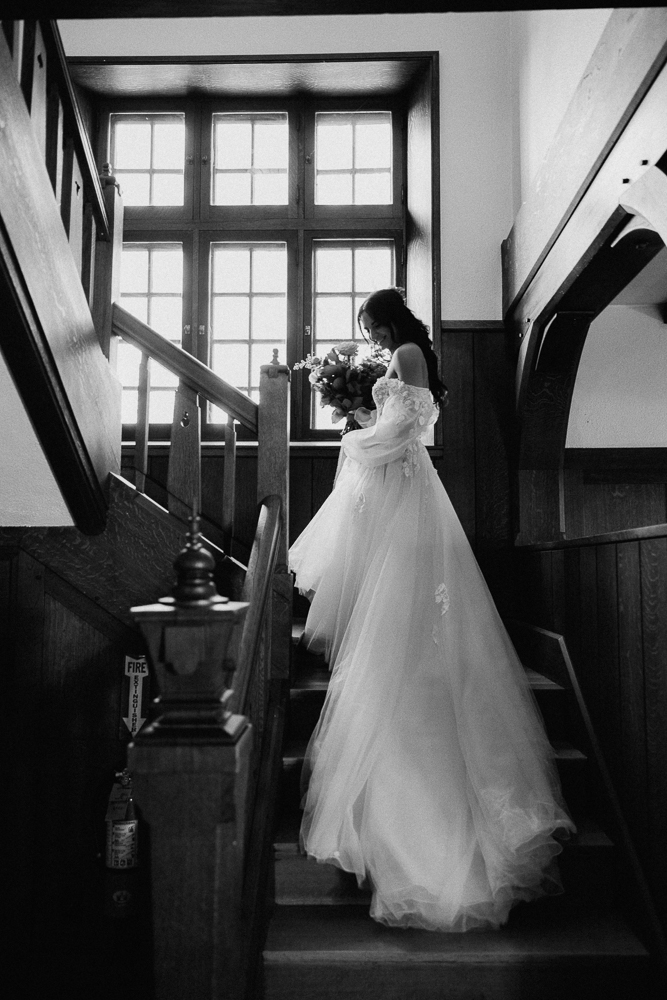 A bride in a flowing wedding gown stands on a wooden staircase, holding a bouquet of flowers, with sunlight streaming through large window panes behind her. The scene is captured in black and white.
