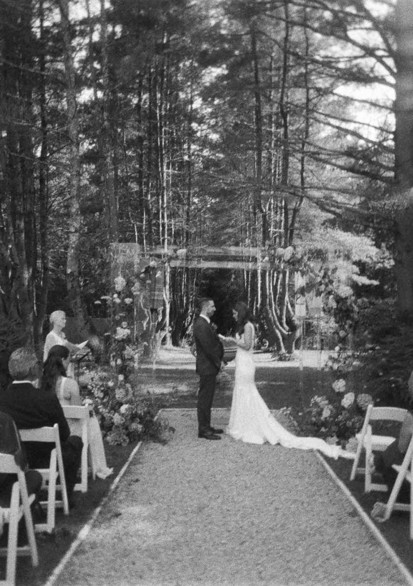 A black-and-white photo of a wedding ceremony outdoors at Seven Hills Inn in Lenox MA, with a bride and groom standing before a floral arch, surrounded by seated guests among tall trees.