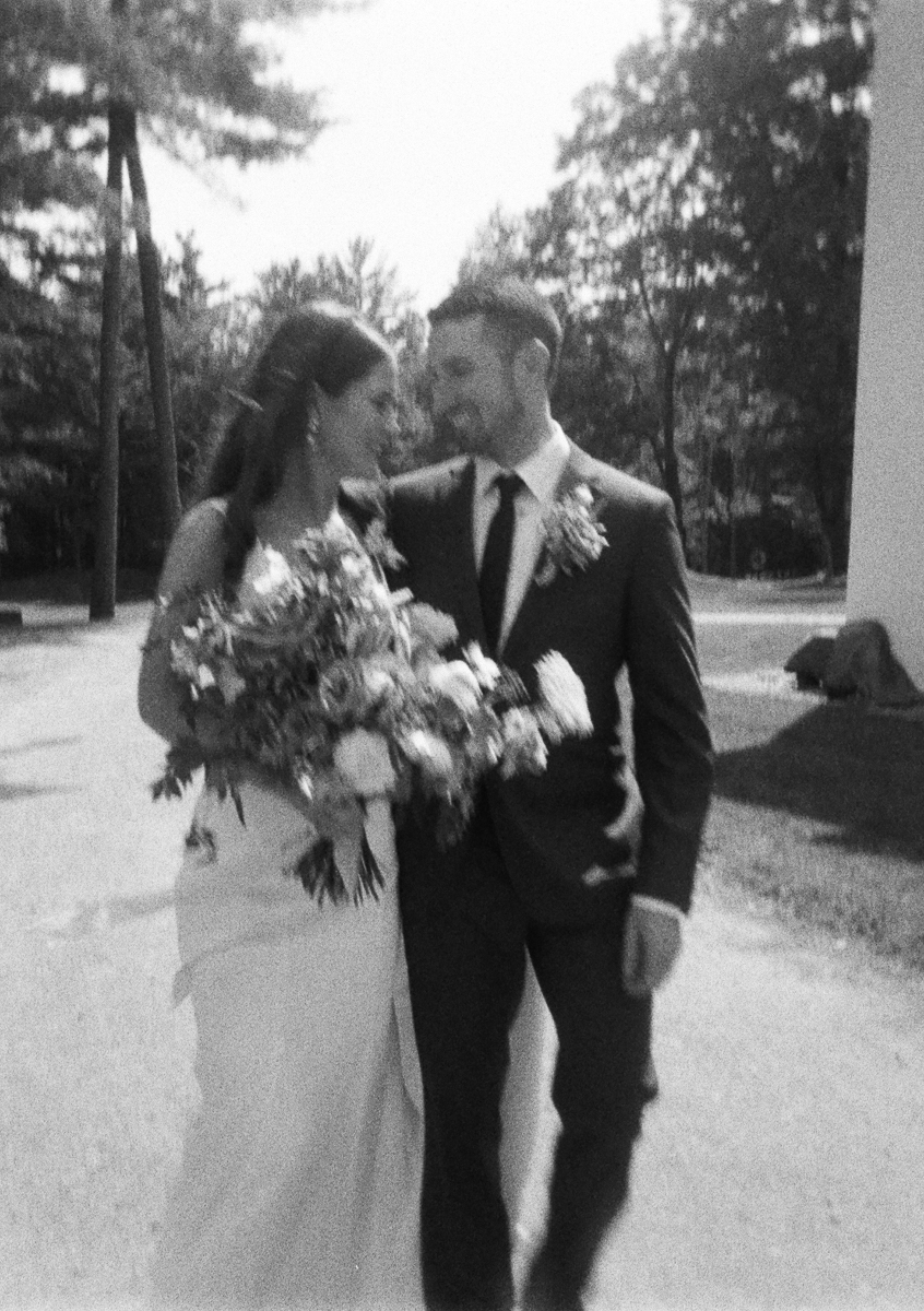 A black and white photo of a bride and groom walking outdoors at their Seven Hills Inn wedding in Lenox MA. Smiling at each other, the happy couple is surrounded by trees and sunlight as the bride holds a large bouquet of flowers.