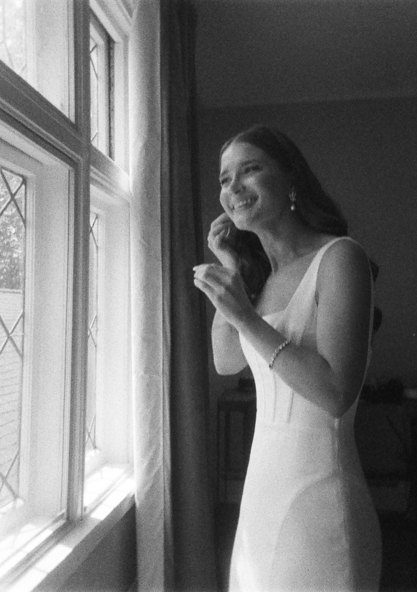 A woman in a white dress smiles as she puts on an earring by a window with patterned glass at Seven Hills Inn in Lenox MA. The black and white photo creates a soft, elegant wedding atmosphere.