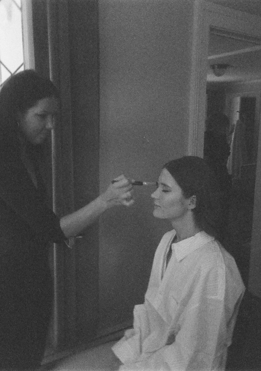 A woman in a white shirt sits while another woman applies makeup to her face with a brush in a dimly lit room, captured in black and white at a Seven Hills Inn wedding in Lenox, MA.