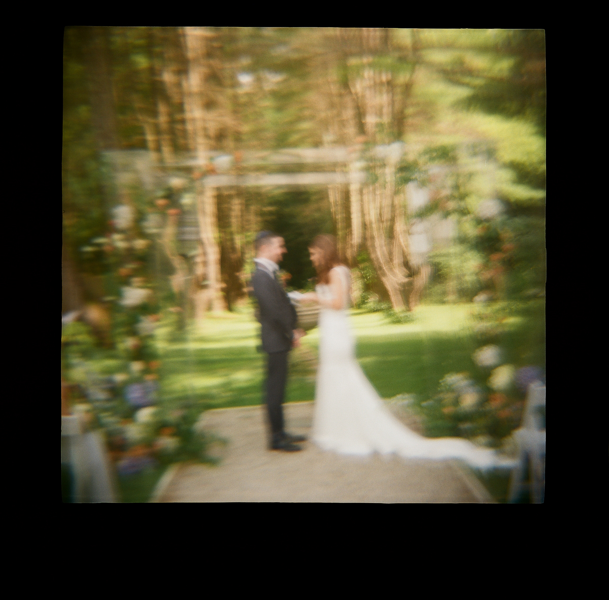 A soft-focus image of a bride and groom standing outdoors at their Seven Hills Inn Wedding in Lenox, MA, holding hands in front of a floral arch with trees and greenery in the background.