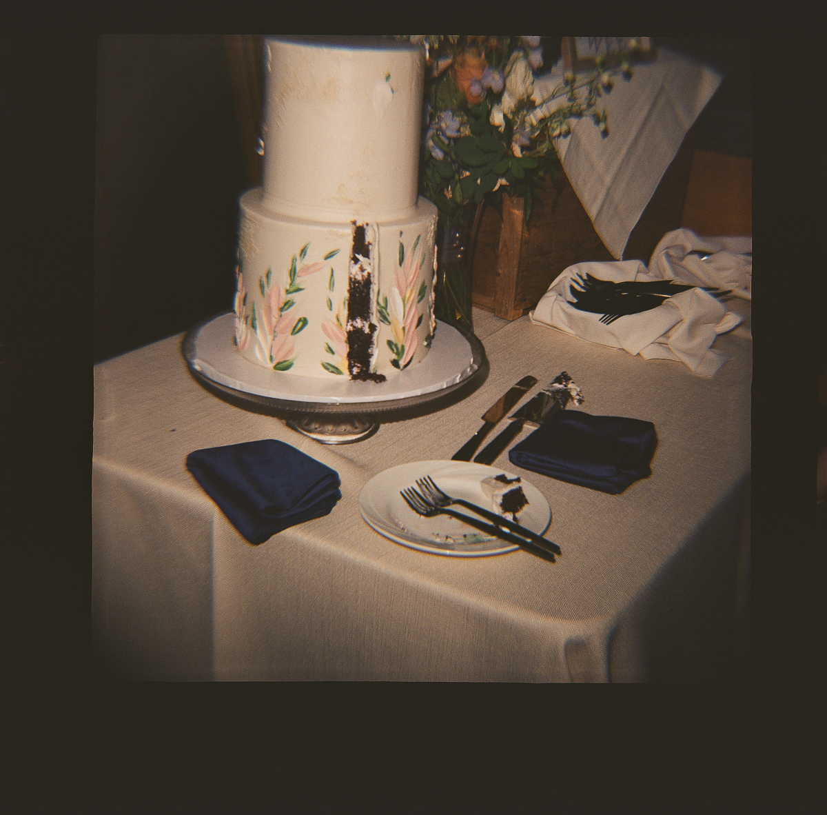 A two-tiered white cake with floral designs sits on a stand at a Seven Hills Inn wedding in Lenox, MA. A slice has been cut out and placed on a small plate with a fork. Blue napkins and utensils are also arranged on the table.