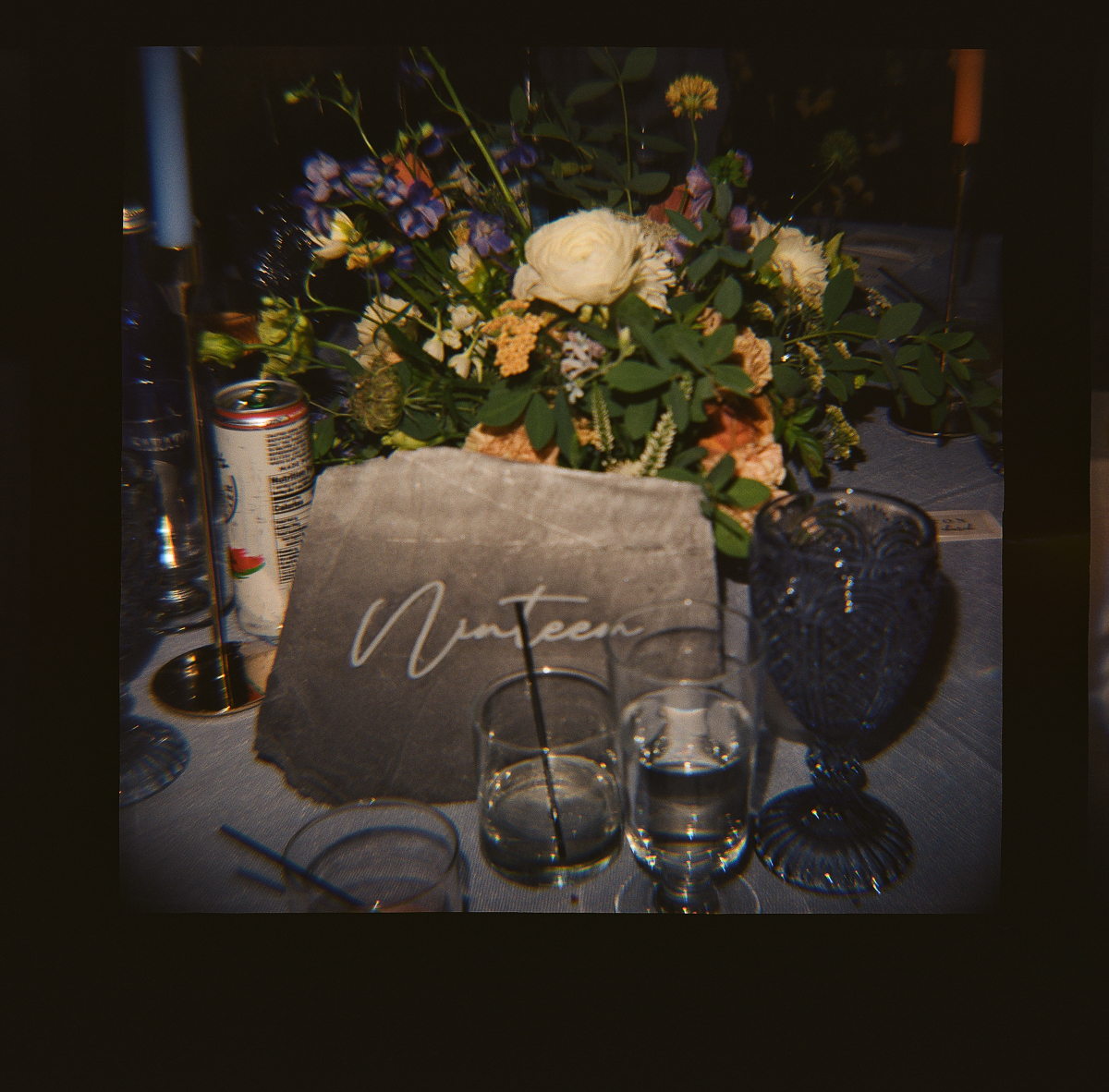 A floral centerpiece sits behind a gray stone with Nineteen written in script at this Lenox MA Seven Hills Inn wedding. The table also has glassware, a can, and a dark blue goblet, all lit by warm, ambient light.