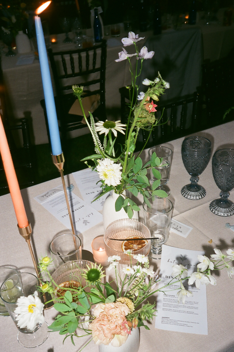 A close-up of a table set for a Seven Hills Inn Wedding in Lenox MA, featuring blue and orange candles, wildflower bouquets in small vases, glassware, and menus on a beige tablecloth. The background reveals more place settings and chairs.