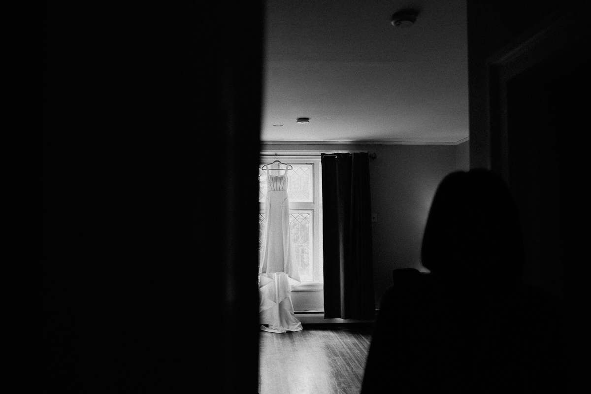A black and white photo shows a wedding dress hanging in front of a window in a softly lit room at Seven Hills Inn, Lenox MA, with a shadowy figure standing in the foreground, partially obscured by darkness.