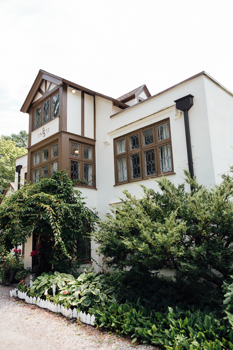 A charming white and brown Tudor-style house in Lenox, MA, with large windows, green vines climbing the walls, and lush garden greenery—perfect for a romantic Seven Hills Inn Wedding on a bright day.