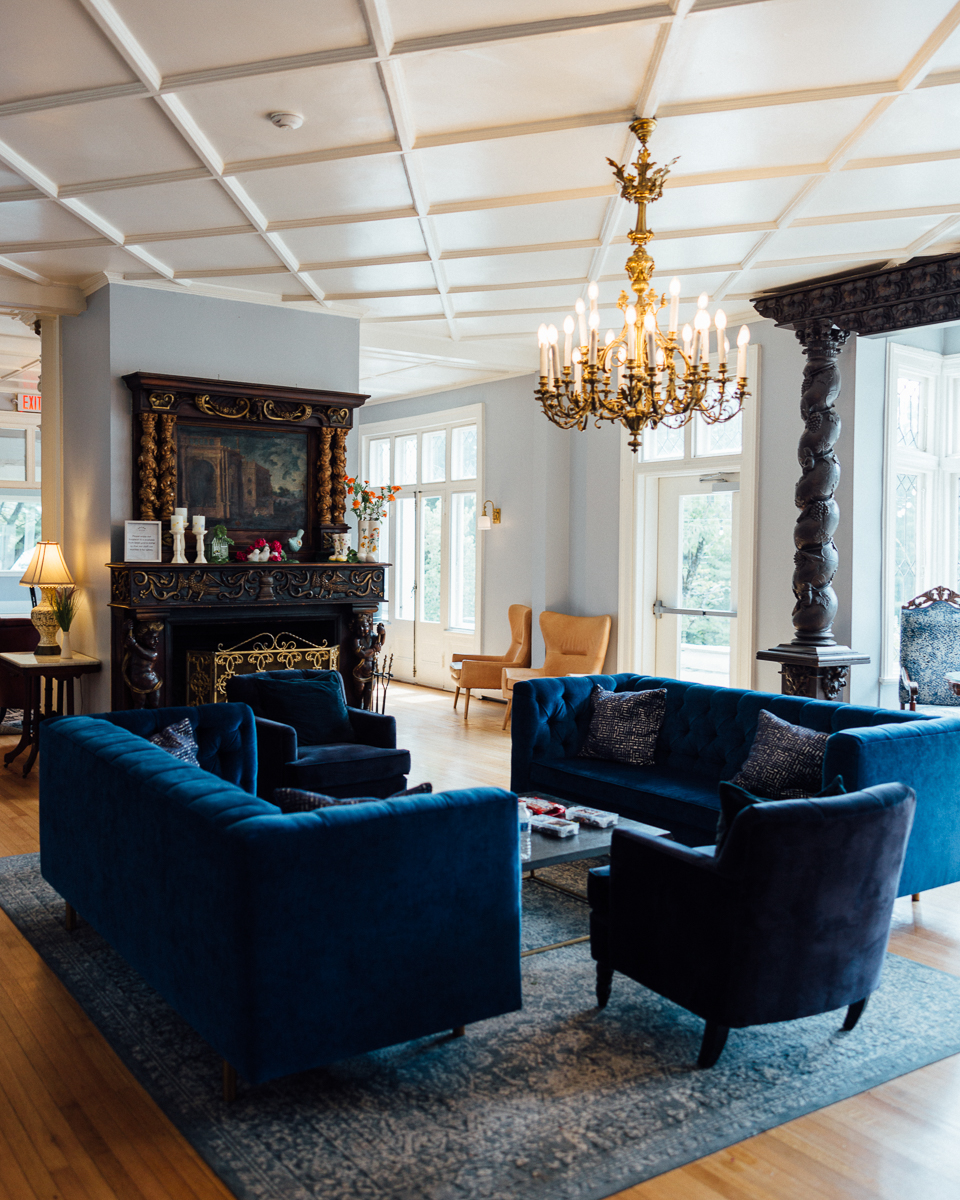 A luxurious living room at Seven Hills Inn, Lenox MA, features blue velvet sofas around a wooden coffee table, an ornate fireplace with mirror, chandelier, large windows, and elegant decor including a rug and carved wooden columns.